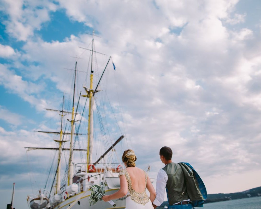 Brautpaar steht vor einem großen Schiff und schaut in den bewölkten Himmel – Symbol für Planung, Wetter und Stimmung bei Schiffshochzeiten